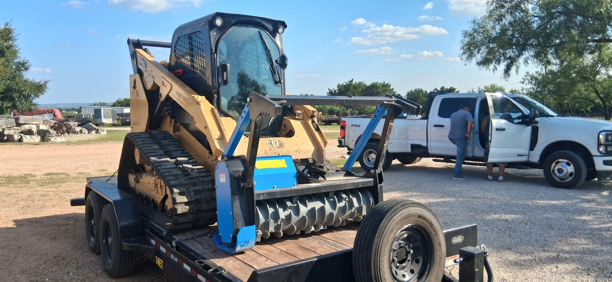 Skid steer loader with blue mulching attachment on flatbed trailer at job site